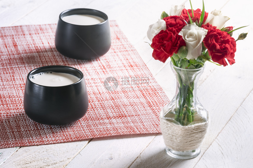 早餐桌两个黑杯子满的鲜奶和一束白玫瑰红康乃馨花在白桌上阳光明媚的清晨图片