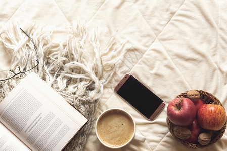 用一本关于羊毛披肩的开放书一个配有苹果和坚的篮子一部电话和杯咖啡的热可背景图片