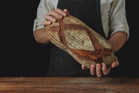 新鲜有机奥瓦尔面包将师的手握在木制桌黑色背景上面包师手握着奥瓦尔面包图片