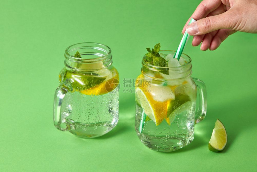 两罐装有天然冷鸡尾酒的玻璃罐子在绿色背景上自制用女手成塑料稻草冷饮酒精或非夏季饮料的概念一名妇女手将天然成分水薄荷柠檬片天然冷自图片
