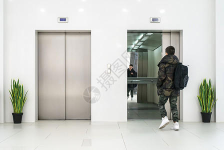 穿便衣和白色运动鞋的少年男孩进入电梯白色当代建筑内部花盆和白色墙中的花朵金属电梯门背景图片