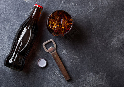 一瓶可乐苏打饮料和玻璃与冰块旁边的开瓶器在黑石上背景图片