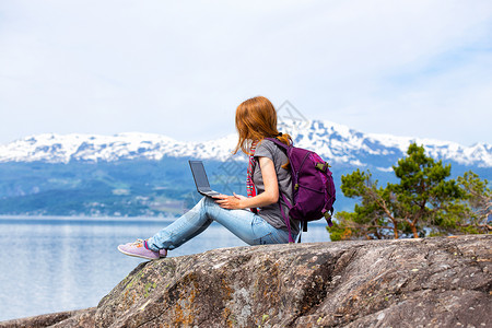 电脑背包穿着笔记本电脑坐在山地和湖泊背景的岩石上背景