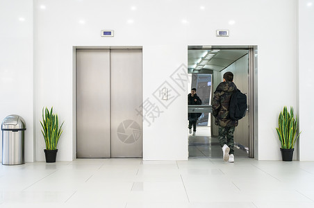 穿便衣和白色运动鞋的少年男孩进入电梯白色当代建筑内部花盆和白色墙中的花朵金属电梯门背景图片