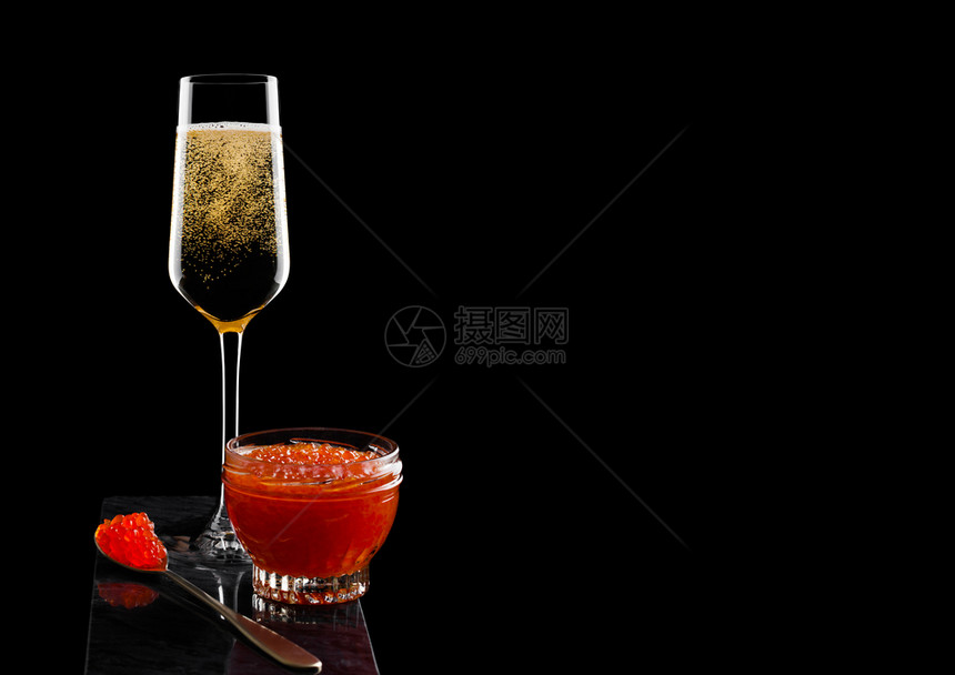 金勺子上配红色鱼酱的优雅黄色香槟杯子和黑底大理石板上装有玻璃容器的鱼子酱图片