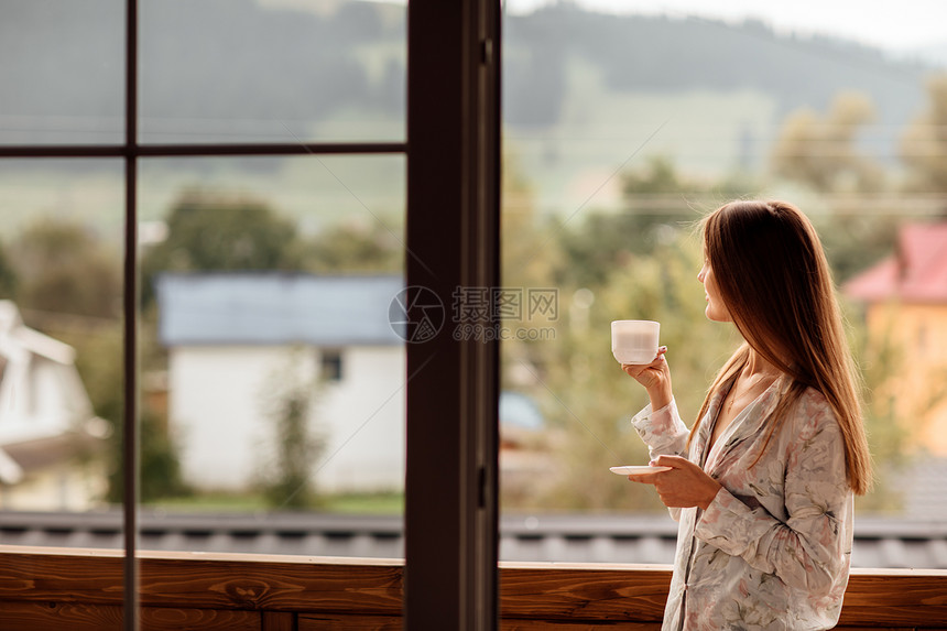 女人早上拿着茶或咖啡看窗边的山图片