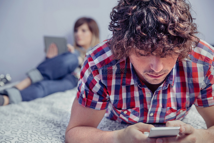 穿着智能手机的年轻英俊男子躺在床上的智能手机和美丽的女人在电子平板上阅读背景在家的休闲时间概念男人在床上使用智能手机女人在平板上图片