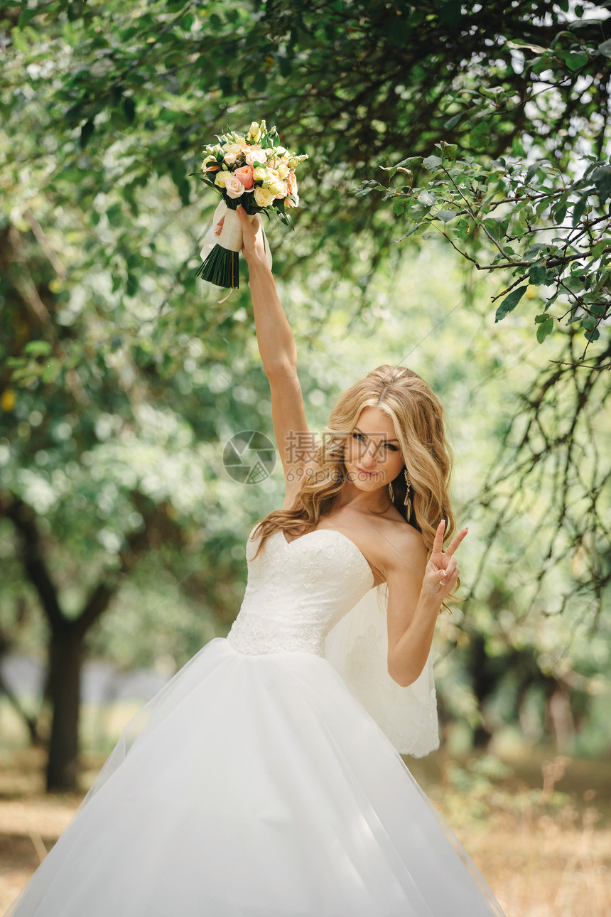 带着长发的可爱年轻新娘拿着她的婚礼花束包括白玫瑰和其他花朵美丽的白色婚纱绿树背景的漂亮女孩长发拿着婚礼花束的可爱年轻新娘白玫瑰和图片