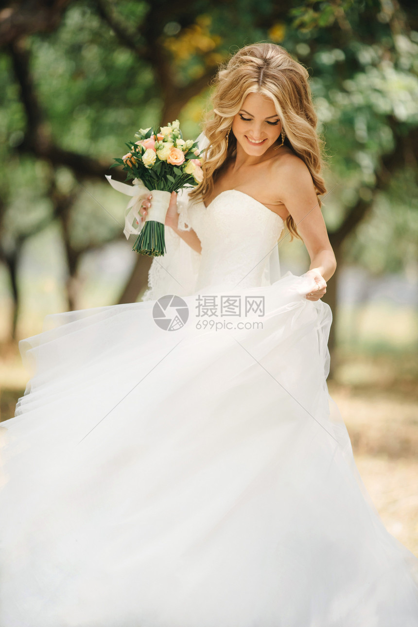 带着长发的可爱年轻新娘拿着婚纱花束包括白玫瑰和其他花朵美丽的白婚礼服绿树背景的漂亮女孩带着长发的可爱年轻新娘拿着婚礼花束的白玫瑰图片