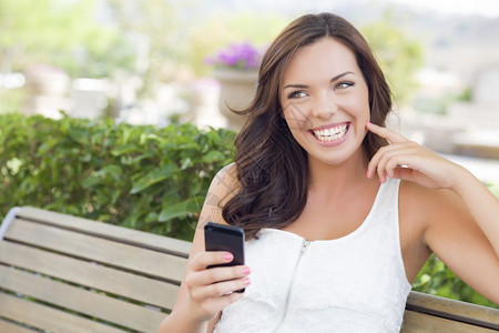 年轻笑的成女在长椅上户外的手机发短信幸福的高清图片素材