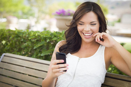 年轻笑的成女在长椅上户外的手机发短信微笑高清图片素材