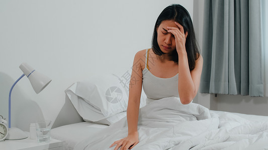 患有不健康疾病的印度裔女患有失眠症身为一名年轻女上午在家卧室的床上吃止痛药以减轻头和喝杯水背景图片