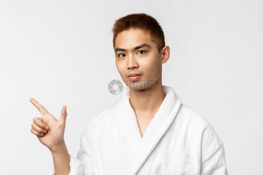美丽spa和休闲概念年轻亚洲男子邀请朋友到温泉的肖像用左手指和看镜头展示广告穿浴袍白色背景美丽spa和休闲概念年轻亚洲男子邀请朋图片
