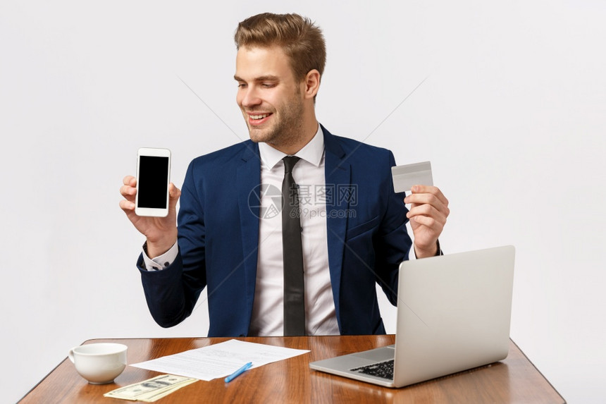 喜悦成功的男创业者在商西装上坐近笔记本电脑咖啡杯和文件持有智能手机和信用卡支付在线购买白背景喜时装和成功的男创业者在商西装上坐近图片