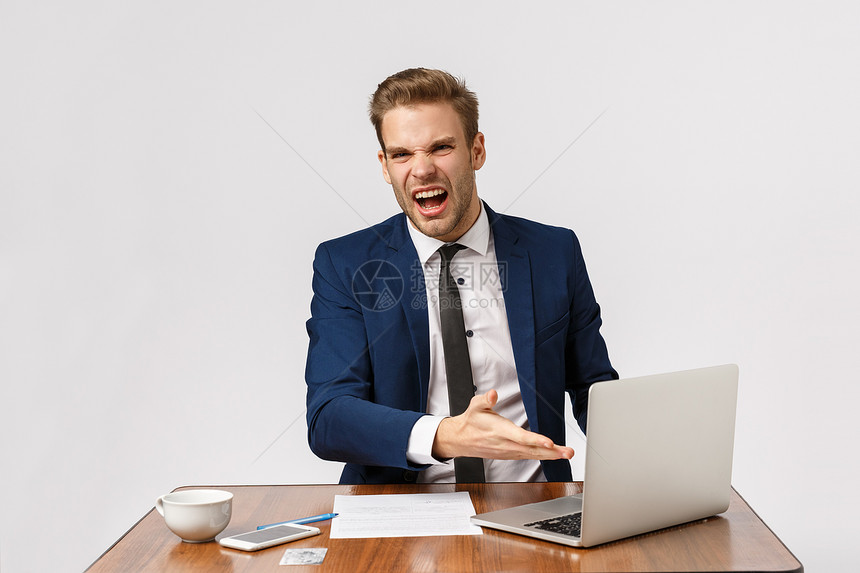 愤怒的年轻老板正式西装的商人坐在办公桌用笔记本电脑显示抱怨的图表站立白色背景这到底是什么真正糟糕的愤怒年轻老板办公桌站在白色背景图片