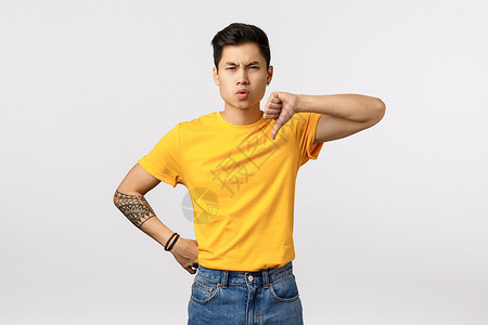 你看起来像个失败者不高兴和失望的年轻英俊亚洲男子纹着黄色t恤衫牛仔裤展示拇指低缩和假人怀疑判断坏的玩耍不赞成你看起来像失败者不满背景图片