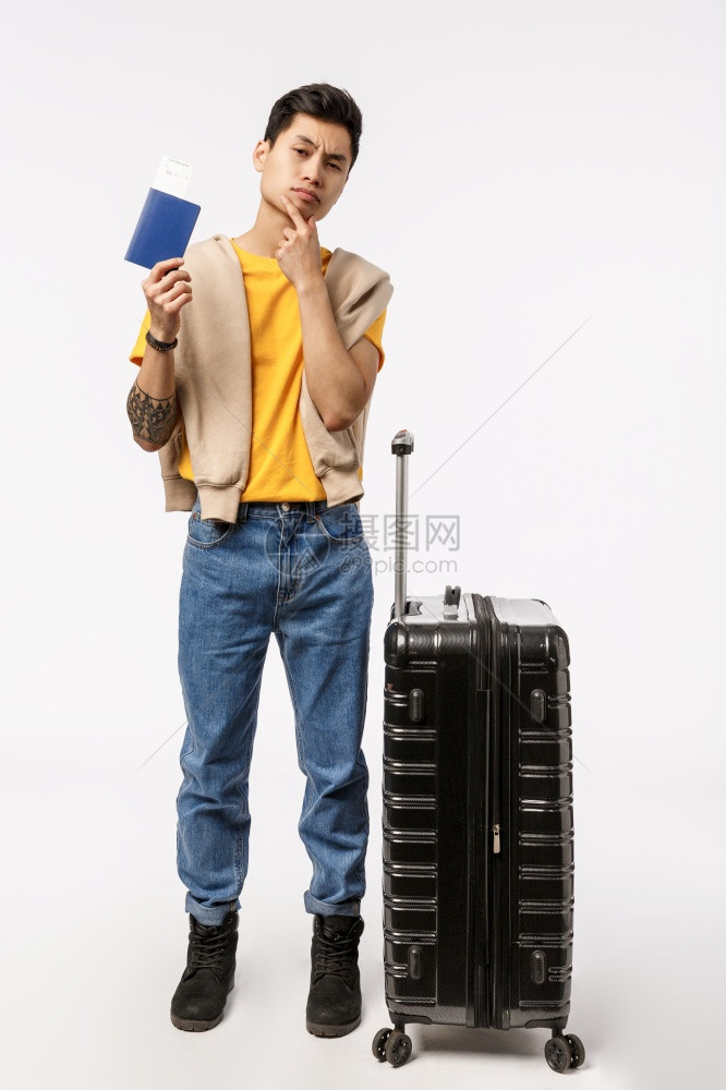 站在黑色手提箱旁边的深思熟虑年轻亚洲男子背包行李决定出国旅学习外摸下巴深思熟虑持有护照购买票纵向全拍照的年轻亚洲男子站在黑色手提图片