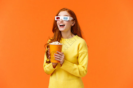 女孩吃爆米花笑的在电影院看大屏幕张开嘴穿着3个眼镜在幻想电影院看站立橙色背景女孩吃爆米花笑的在电影院看大屏幕电影站立橙色背景图片