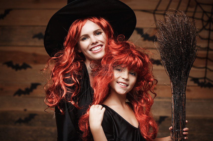 神圣的概念美丽天主教母亲和女儿穿着长红头发的服庆祝万圣节在木制工作室背景上披蝙蝠和蜘蛛网美丽的天主教母亲和女儿穿着长红头发的服庆图片