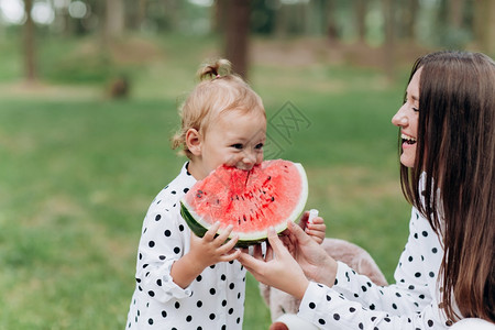 抱西瓜的小孩快乐的母亲和女儿在夏季公园吃西瓜快乐的笑脸家庭在公园吃西瓜母亲和女儿一起度过时间饮食维生素健康的食物概念选择焦点饮食健康的物概念背景