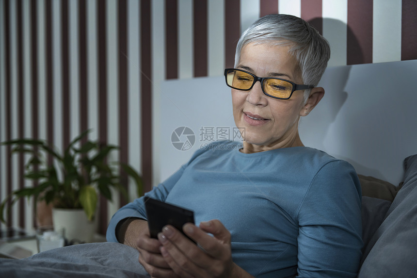 睡前用智能手机把眼镜放在床上晚用智能手机把眼镜用蓝色灯挡住的老年妇女图片