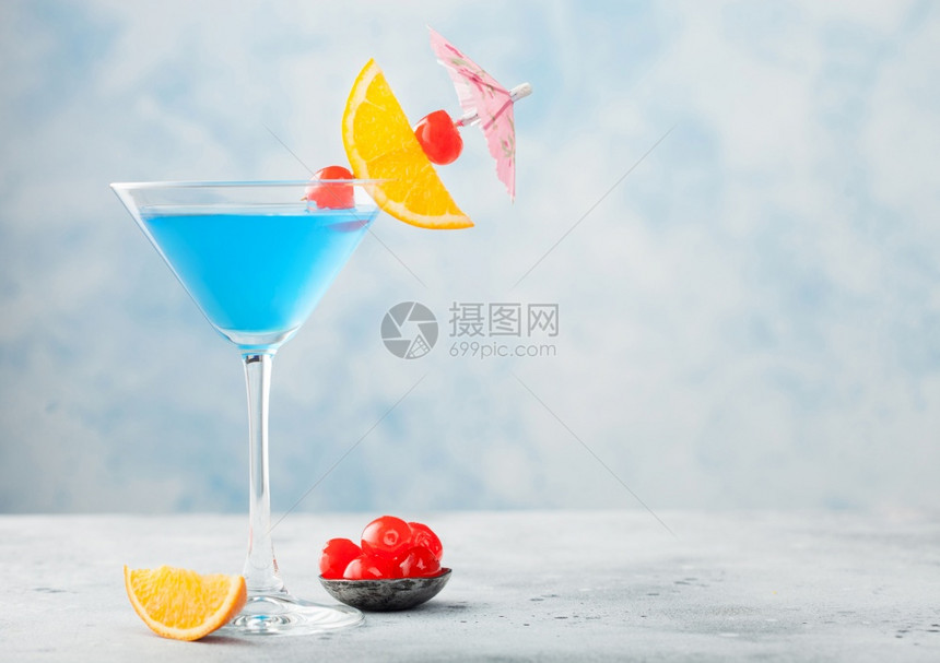 玛蒂尼杯中的蓝色环礁湖夏季鸡尾酒配有甜樱桃和橙色切片蓝桌底有雨伞图片