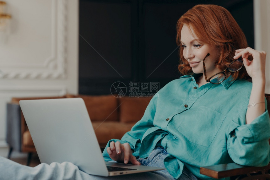 喜悦的年轻红发妇女坐在舒适的扶手椅上做远程工作在社交网络上读新闻忙于电脑工作集中在笔记本电脑的监控上图片