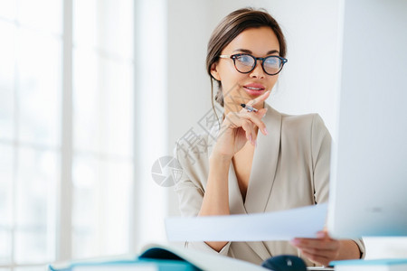 女经济学家在工作空间担任职务侧重于监测核实或对财务报告中的数字持有纸和笔染黑头发穿正式商业西装背景图片