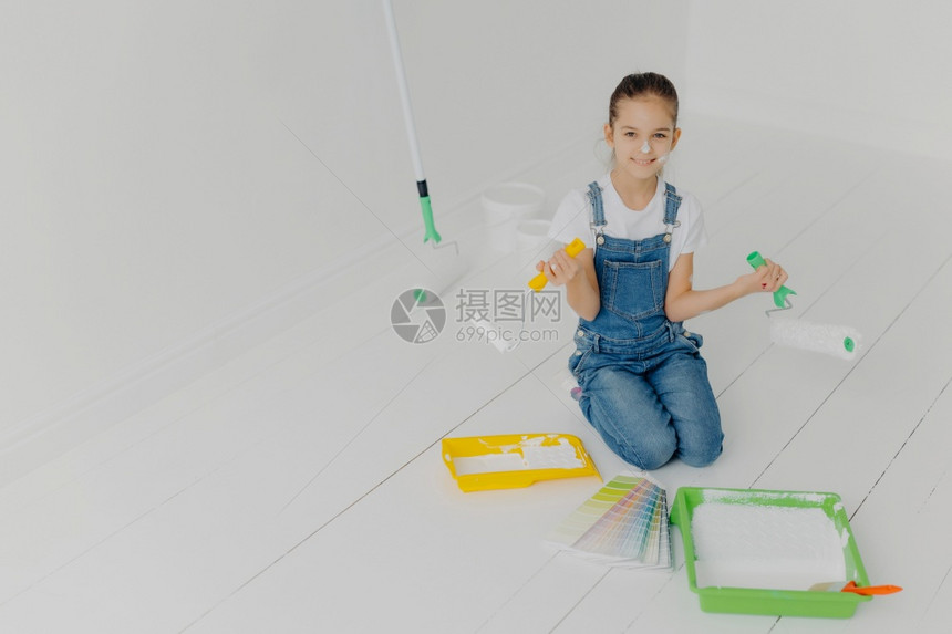 戴尼姆服饰中的欧洲小女孩顶部观视跪下用油漆滚机帮助父母改造房子在地板上摆姿势在样本中选择颜色有快乐的表情图片