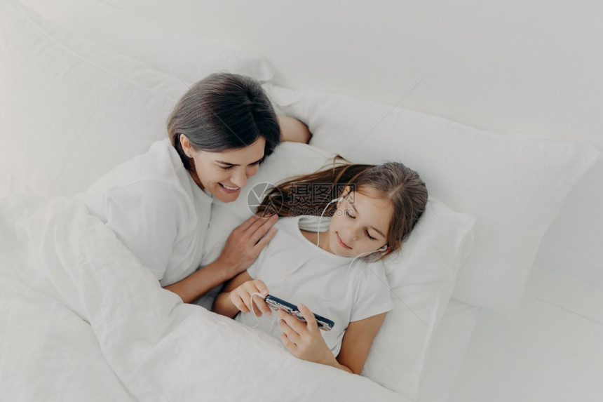 快乐的妈被拍到幸福母亲与可爱的小女儿共度空闲时间她专注于智能手机听耳的歌穿着白色软睡衣躺在床上周末早懒惰图片