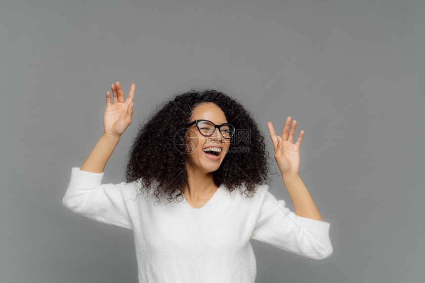快乐的美国女人举起手来快乐地笑享受愉快的音乐戴光学眼镜穿白色的随意跳跃者模范在灰色背景之上复制面的文字空间图片