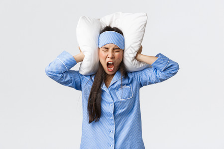 愤怒的亚洲女孩穿着睡衣和面具躺在床上用枕头闭耳朵大声叫喊因为吵得睡不着邻居在晚上通过聚会抱怨烦人的声音愤怒亚洲女孩穿着睡衣和面具背景图片
