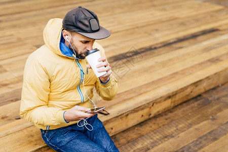 穿着大胡子戴帽黄外套和牛仔裤的时装男子肖像喝着美味咖啡图片