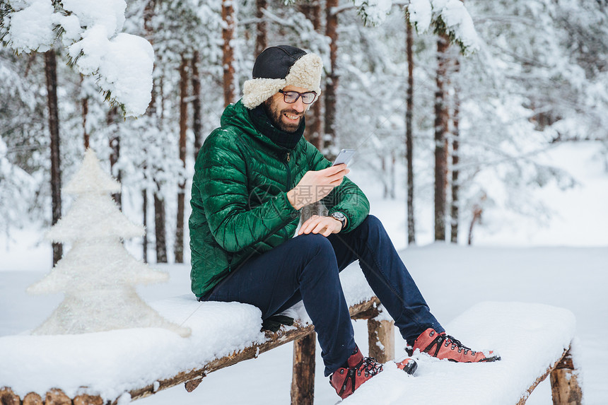 男在冬季穿着温暖的衣在手机上阅读信息在冬季森林户外的平静气氛中自由度过时间享受新鲜空气人休息技术概念图片