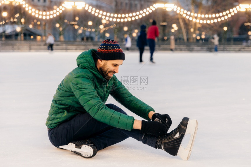 帅哥穿绿色外套和帽子坐在冰上和鞋带滑冰快乐的表情帅哥在滑冰圈上过冬假季和节概念图片
