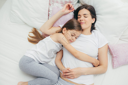 做个好梦快乐的困睡母亲和可爱的女儿在床上拥抱做个愉快的梦躺在白睡衣上有良好的关系互相照顾背景