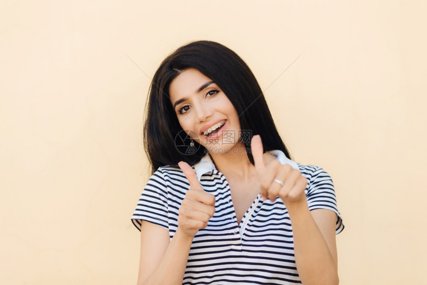 高兴的年轻女士举起拇指赞同一些乐观的表达方式穿着随意的条纹T恤衫与工作室背景隔绝图片