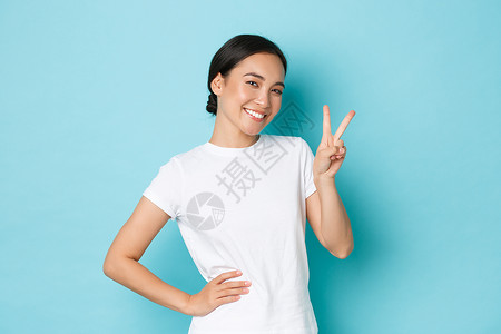穿着白色t恤衫的可爱笑亚洲女展示和平kawi标志和看起来可爱调情保持积极或乐观站立蓝背景的快乐态度背景图片