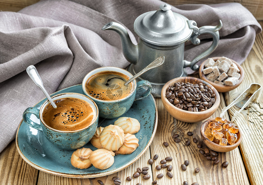 两杯蓝色黑咖啡饼干和一锅咖啡上面包着旧木制桌布甘蔗糖和咖啡豆图片
