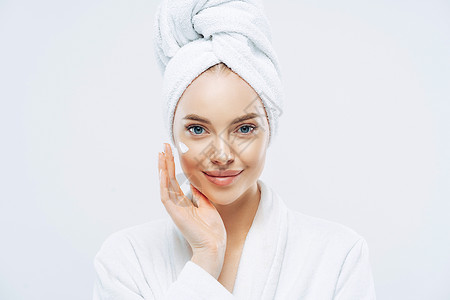 年轻自信的causin女人使用面霜享受新的反皱纹化妆品产防止皮肤衰老的迹象穿最小化妆品穿着浴袍隔离在白墙上背景图片