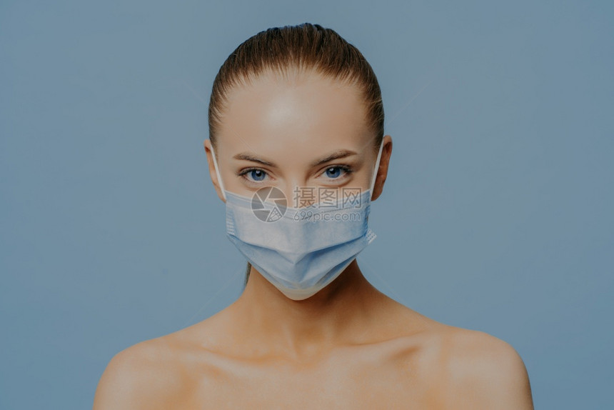 青壮的年轻女模型直视照相机肩膀赤身室内穿戴医疗面具以保护蓝底隔离的冠状图片