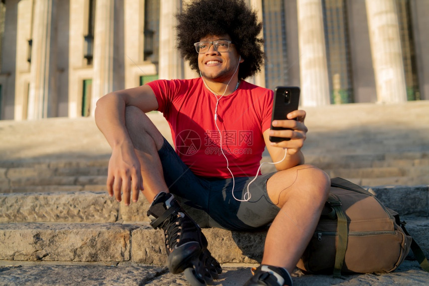 年轻拉丁男子使用手机的肖像坐在户外时穿滑雪鞋体育概念城市图片
