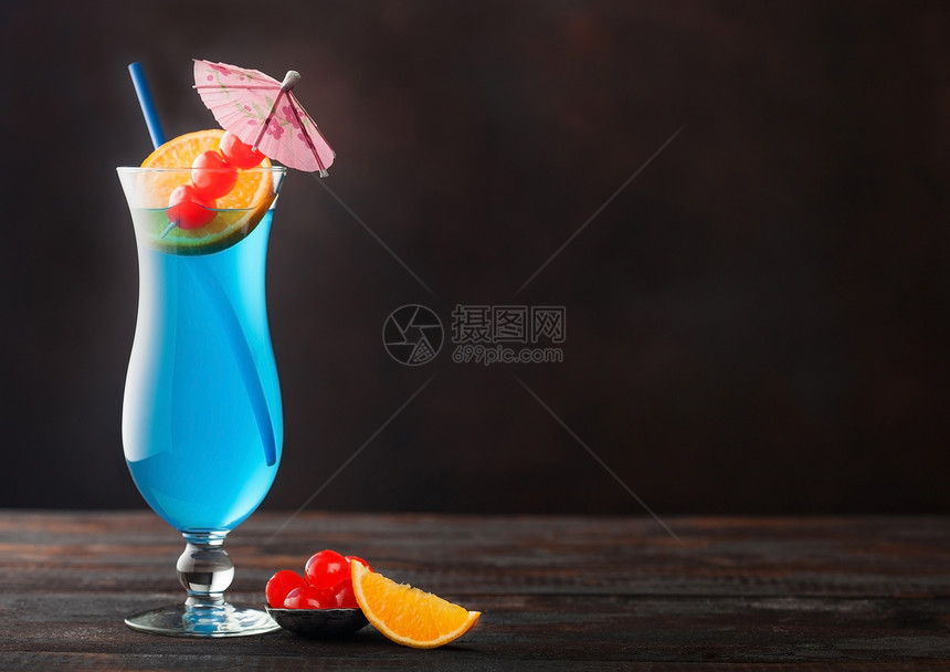 经典玻璃中的蓝色环礁湖夏季鸡尾酒有甜樱桃和橙色切片黑桌底有雨伞图片