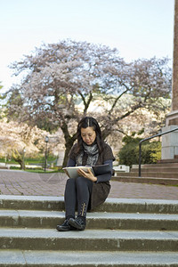 亚裔妇女坐在台阶上同时持有笔记本垫底有樱桃树和蓝天空图片