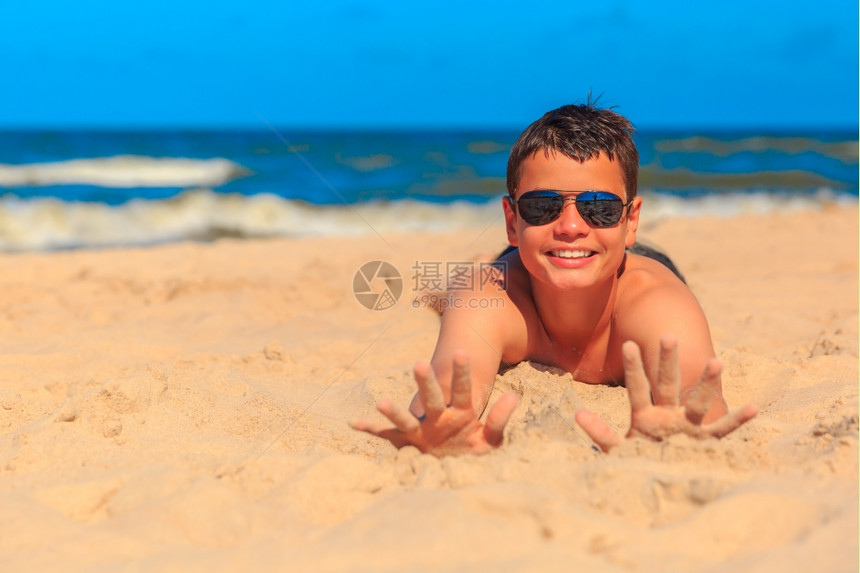13岁快乐的笑男孩在海边展示了沙中的双手浅田地深度专注在手和脸上图片