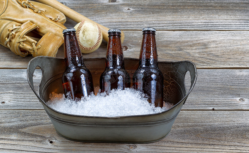 三瓶棕色装啤酒金属桶中压碎的冰块和生锈木上背景的棒球设备前方三股褐色瓶装啤酒金属桶中压碎的冰和粗木上背景的棒球设备图片