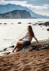 美丽的年轻女坐在海滩上的大石头看着遥远的岛屿图片