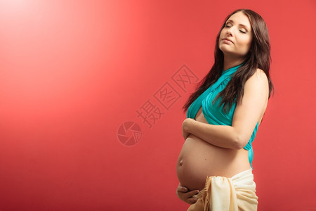 怀孕母亲和幸福概念穿着围巾的有吸引力孕妇在红色背景下触摸肚子室内的高清图片素材