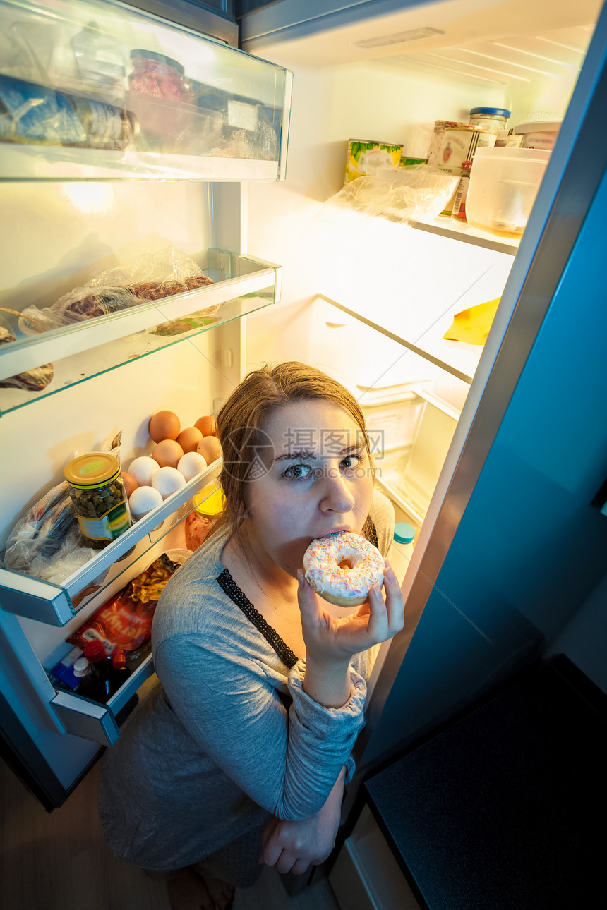 穿着睡衣在冰箱旁边吃甜圈的年轻妇女肖像图片