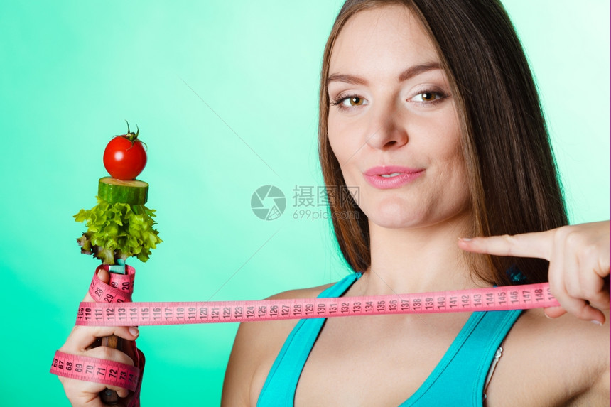健美的身女拿着新鲜混合蔬菜的叉子和绿色蓝背景的测量胶带图片